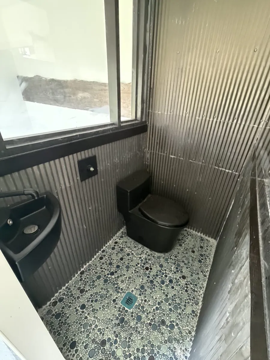 Compact wet bath inside the 20-foot mobile container with black fixtures, corrugated wall panels, and pebble-look shower flooring