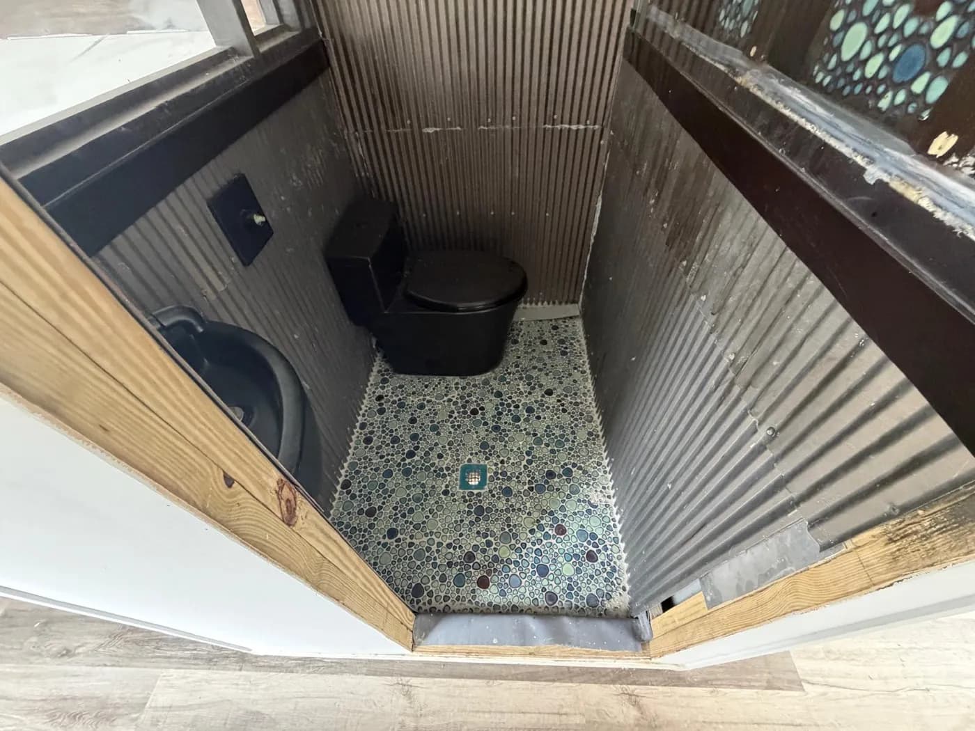 Private bathroom inside the container mini-home with corrugated metal walls, a black toilet, and a pebble-tile floor