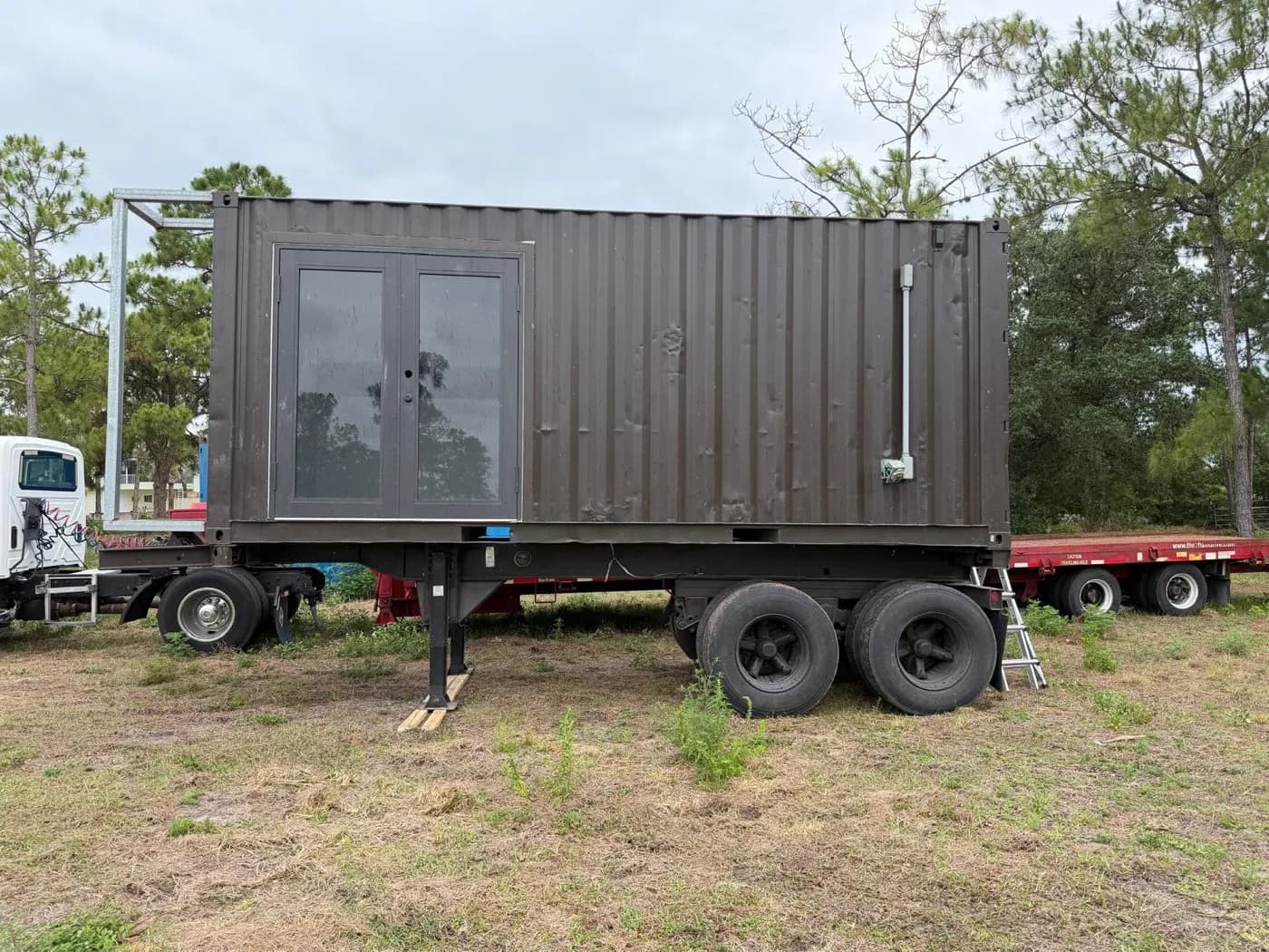 Charcoal-painted container mini-home on a tandem-axle trailer with full-glass French doors, parked on a grassy Southwest Florida lot
