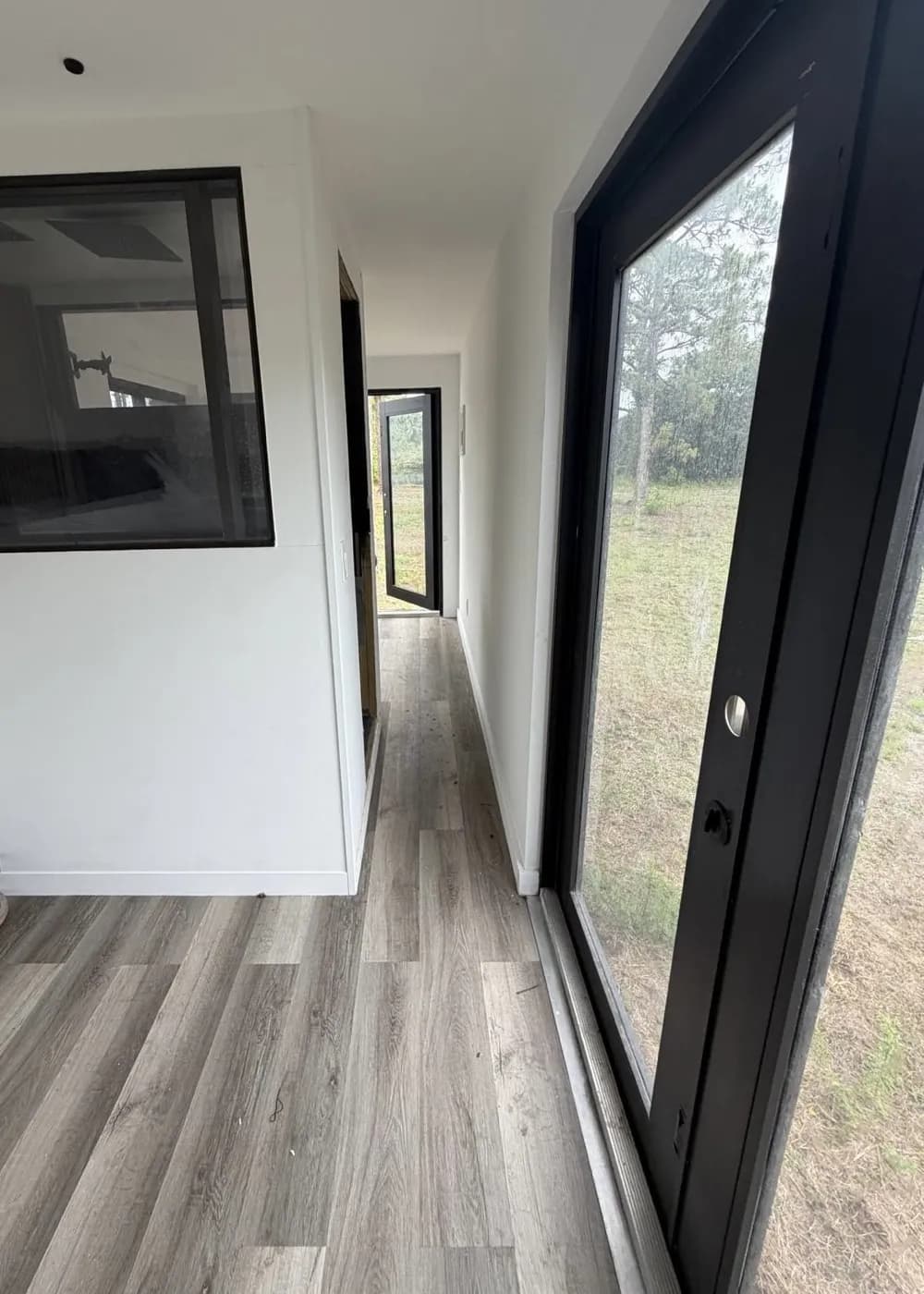 Interior of the container mini-home looking down the hallway toward a full-glass entry door, with gray wood-look vinyl plank flooring and white walls