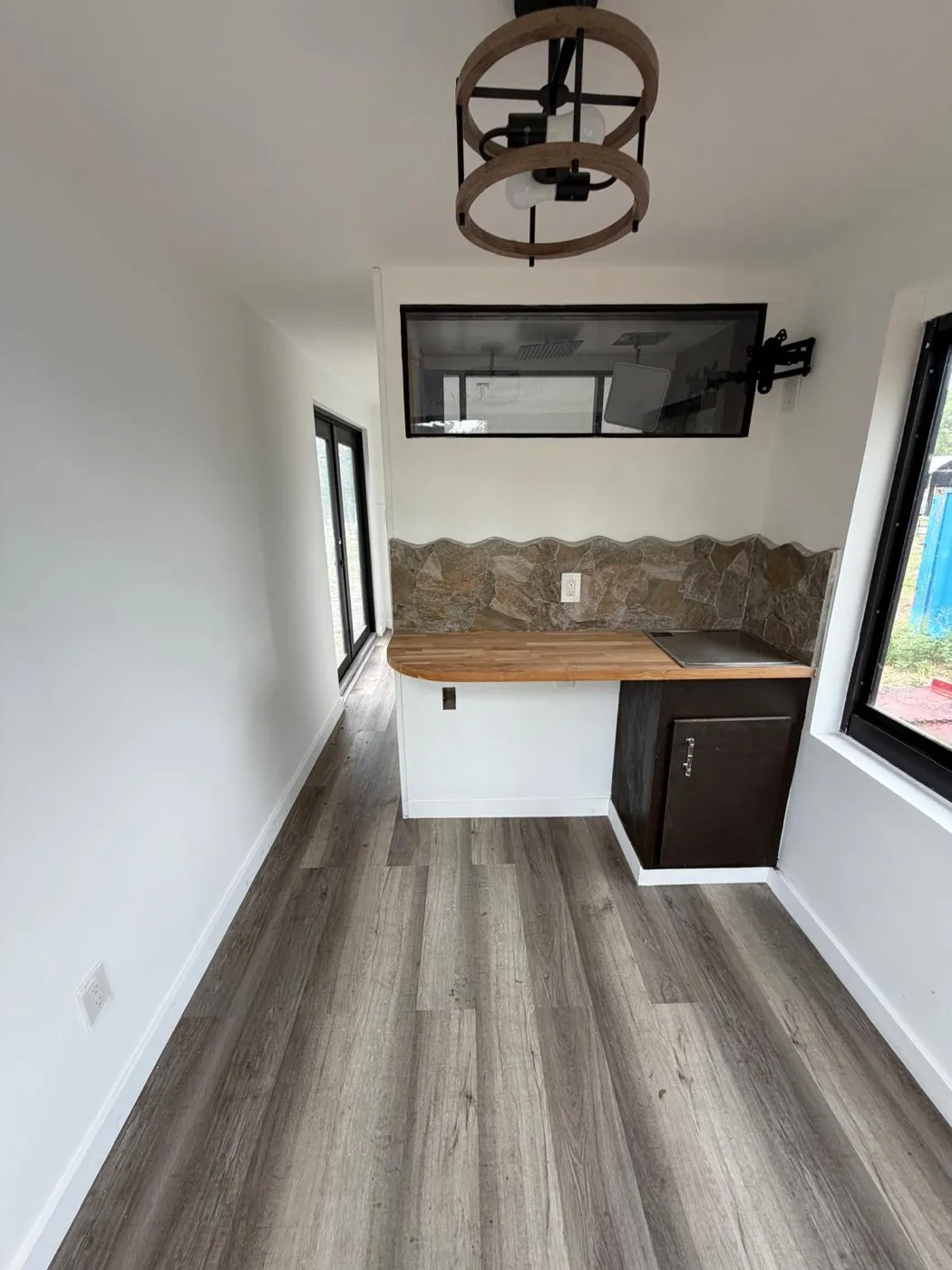 Container mini-home kitchenette with butcher-block countertop, natural stone backsplash, cooktop, dark base cabinet, and a wagon-wheel light fixture